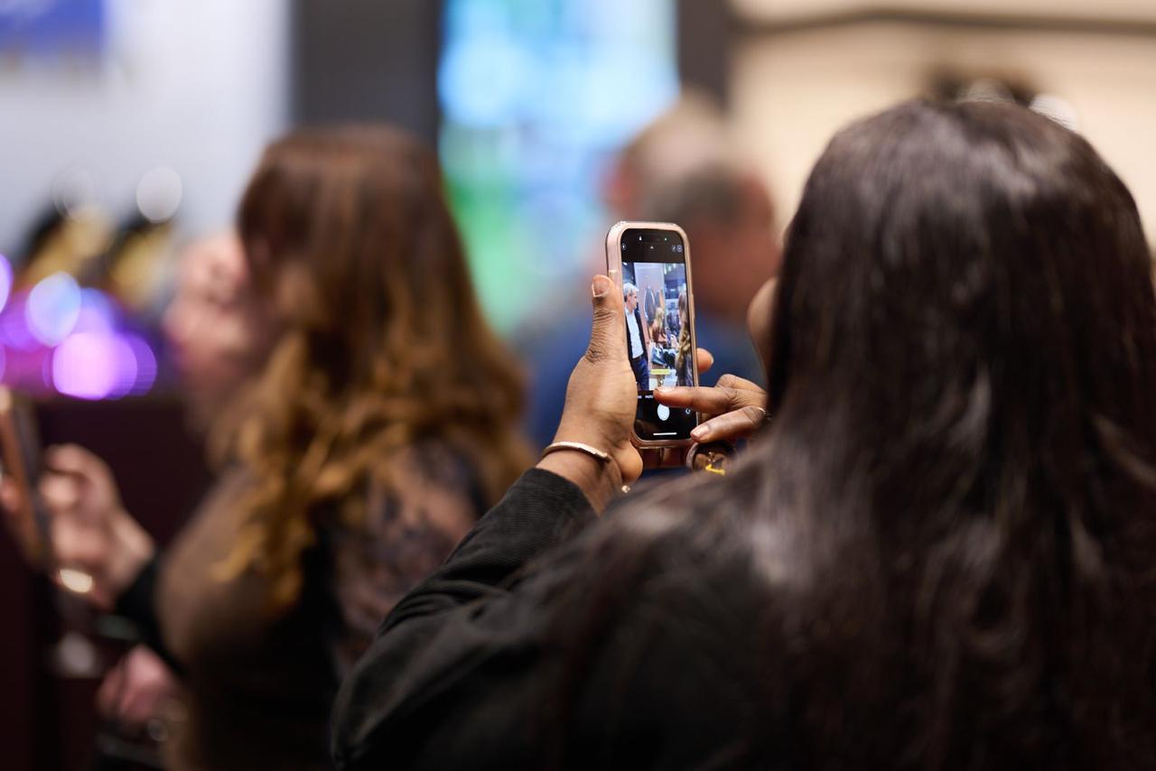 Tofunmi Balogun from behind, phone raised, capturing a room full of people